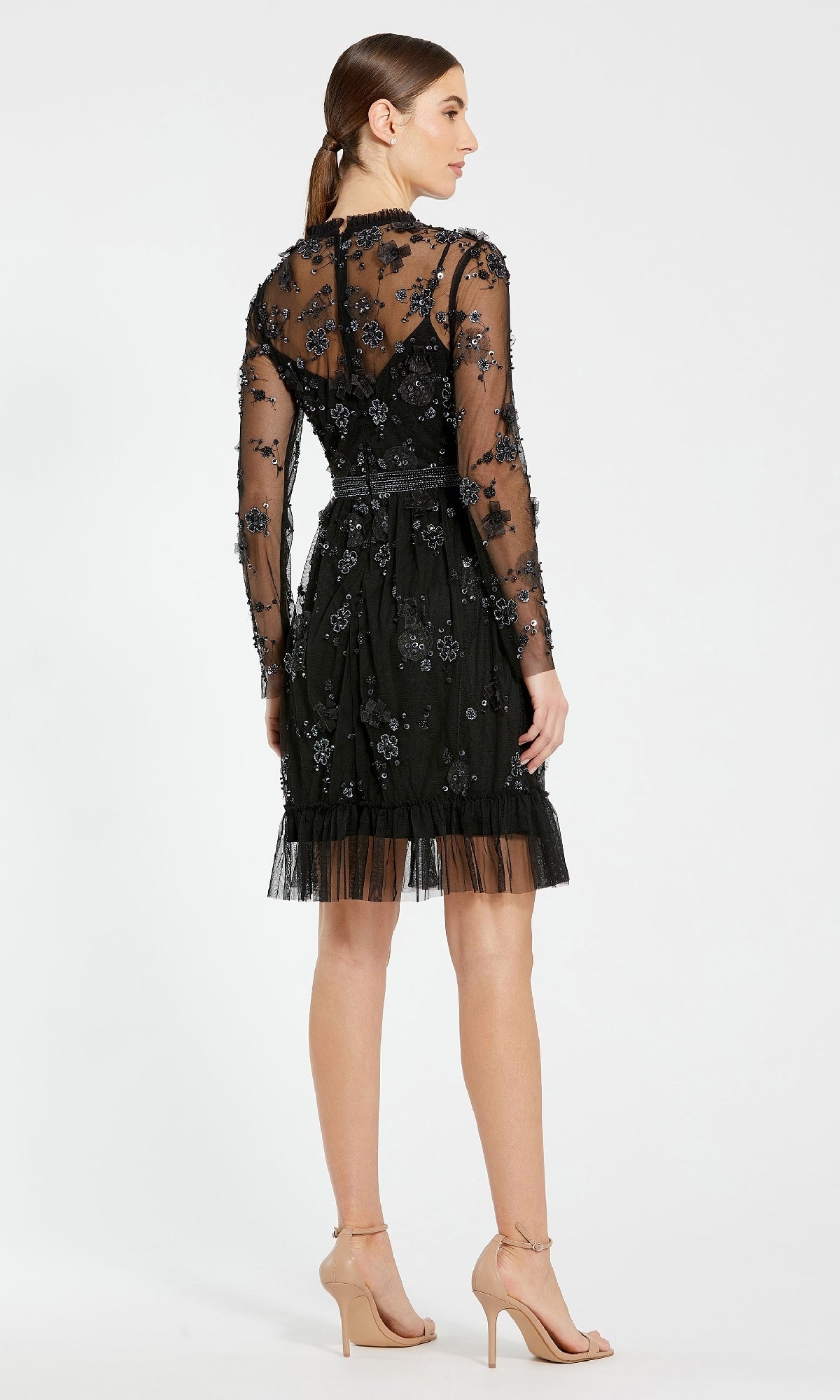 A woman stands in profile wearing the Mac Duggal 9140, a short black beaded dress with floral embellishments—an elegant wedding guest choice—paired with nude high heels on a plain background.