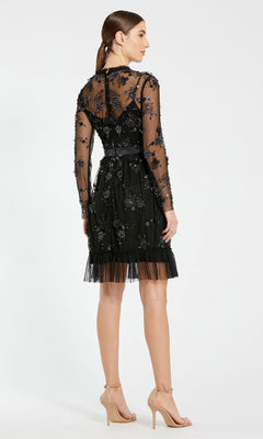 A woman stands in profile wearing the Mac Duggal 9140, a short black beaded dress with floral embellishments—an elegant wedding guest choice—paired with nude high heels on a plain background.