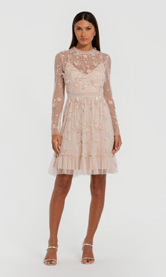 A woman wears the Mac Duggal 9140 short blush wedding guest dress featuring long sleeves, sheer nude fabric, floral embroidery, tulle skirt, and pearl details, paired with strappy heeled sandals.