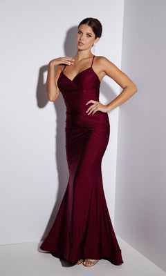 A woman stands in a corner wearing the Classic Long Mermaid Prom Dress: Eureka 9151, a fitted, floor-length burgundy gown with spaghetti straps—perfect for any formal occasion.