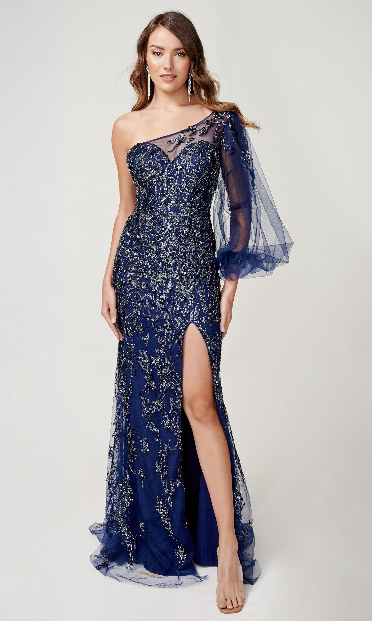A woman models the Nina Canacci 9159 long formal dress in navy blue, featuring a one-shoulder neckline, beaded details, a sheer sleeve, and a thigh-high slit against a plain background.
