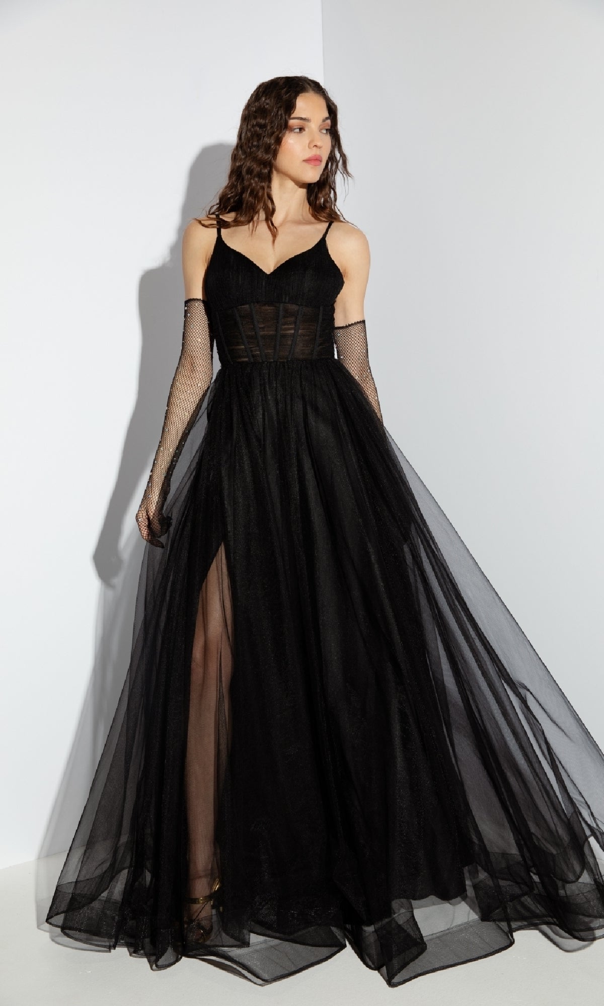 A woman stands against a white background wearing the Corset Long Tulle Prom Ball Gown: Eureka 9177, featuring a sheer corset bodice, long black skirt with a thigh-high slit, and paired with long mesh gloves.