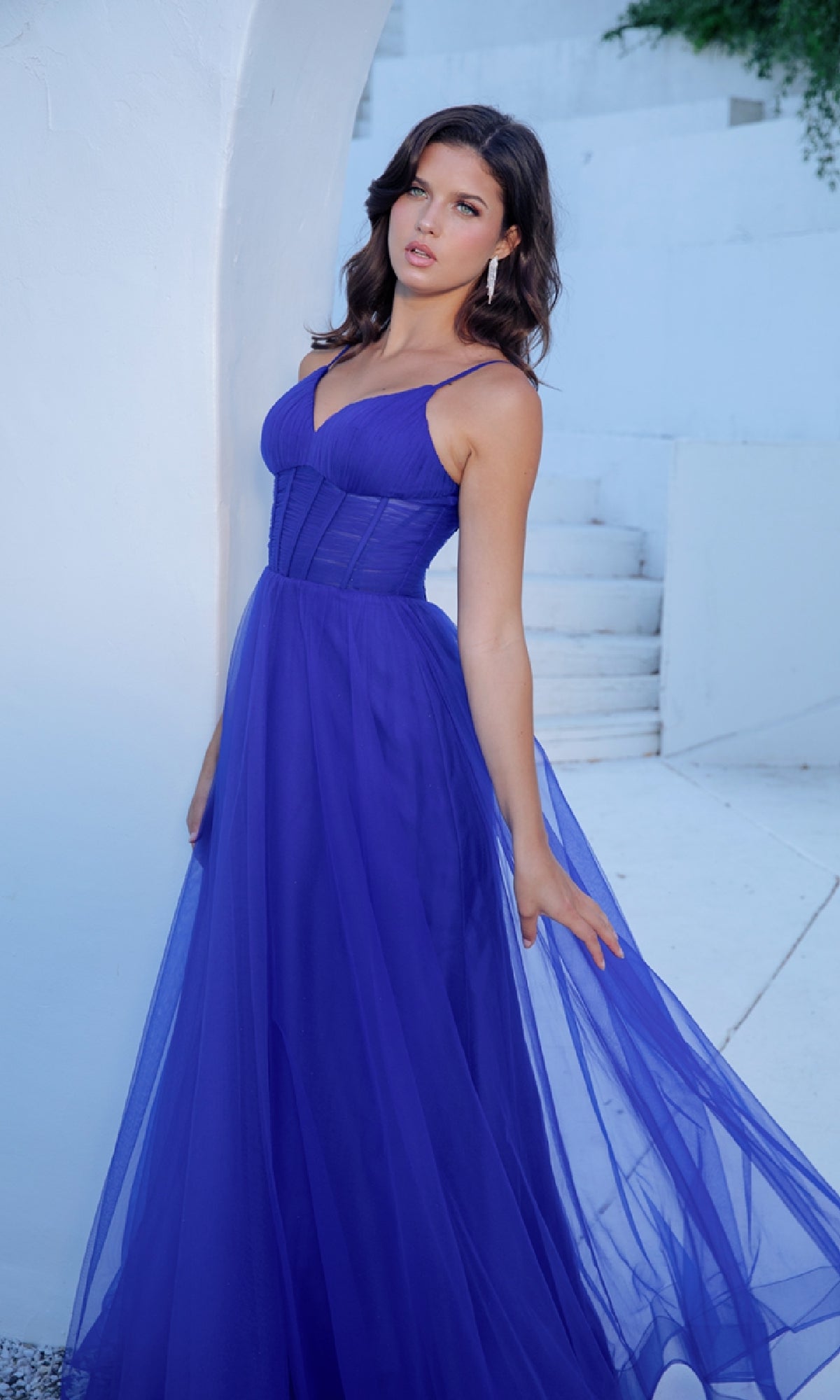 A woman poses outdoors in the Eureka 9177 Corset Long Tulle Prom Ball Gown, a royal blue dress with a fitted corset bodice and sheer, floor-length skirt. White stairs and walls provide a chic backdrop.