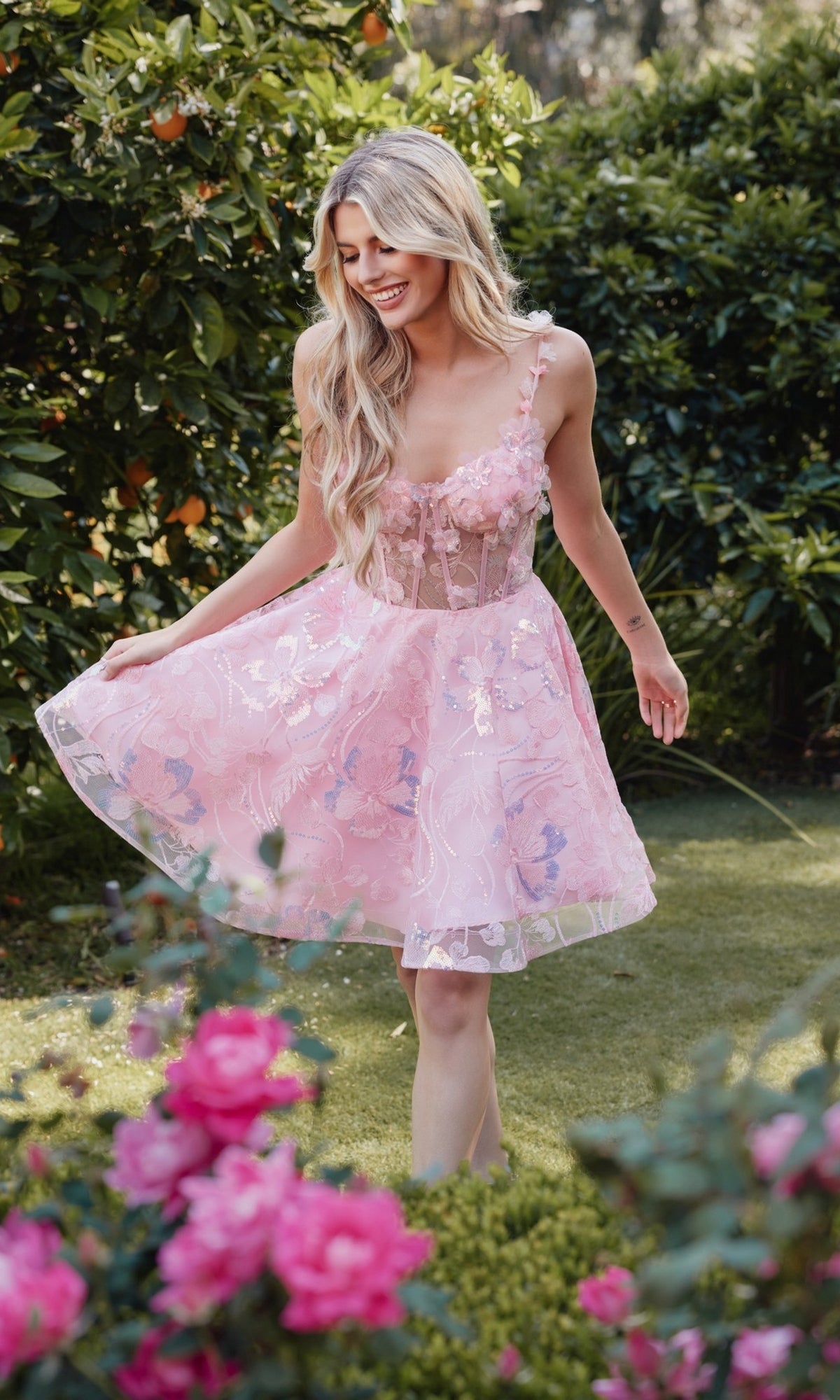 A woman with long blonde hair stands on grass in a garden, surrounded by green bushes and blooming pink flowers, wearing the Sheer-Corset Short Butterfly Hoco Dress JT917A.