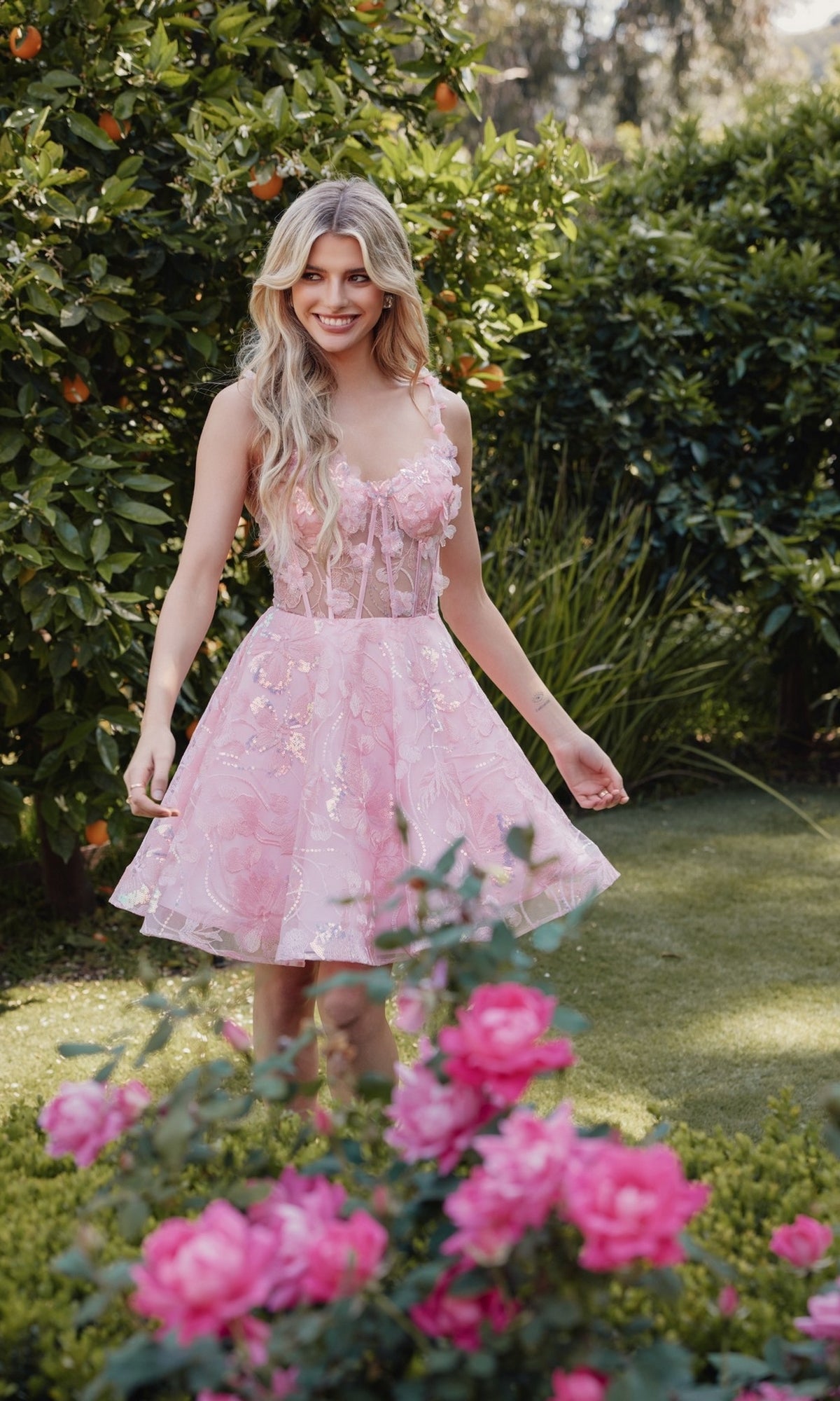 A woman wearing the Sheer-Corset Short Butterfly Hoco Dress JT917A smiles at the camera while standing in a garden with green bushes and pink flowers.