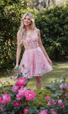 A woman smiles in a garden, standing on grass among blooming pink flowers and green foliage, wearing the Sheer-Corset Short Butterfly Hoco Dress JT917A.