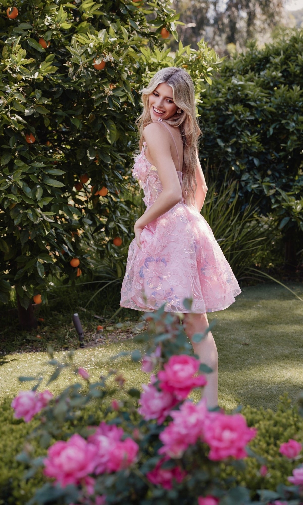 A woman in the Sheer-Corset Short Butterfly Hoco Dress JT917A stands on grass in a garden, smiling over her shoulder amid orange trees and pink flowers.