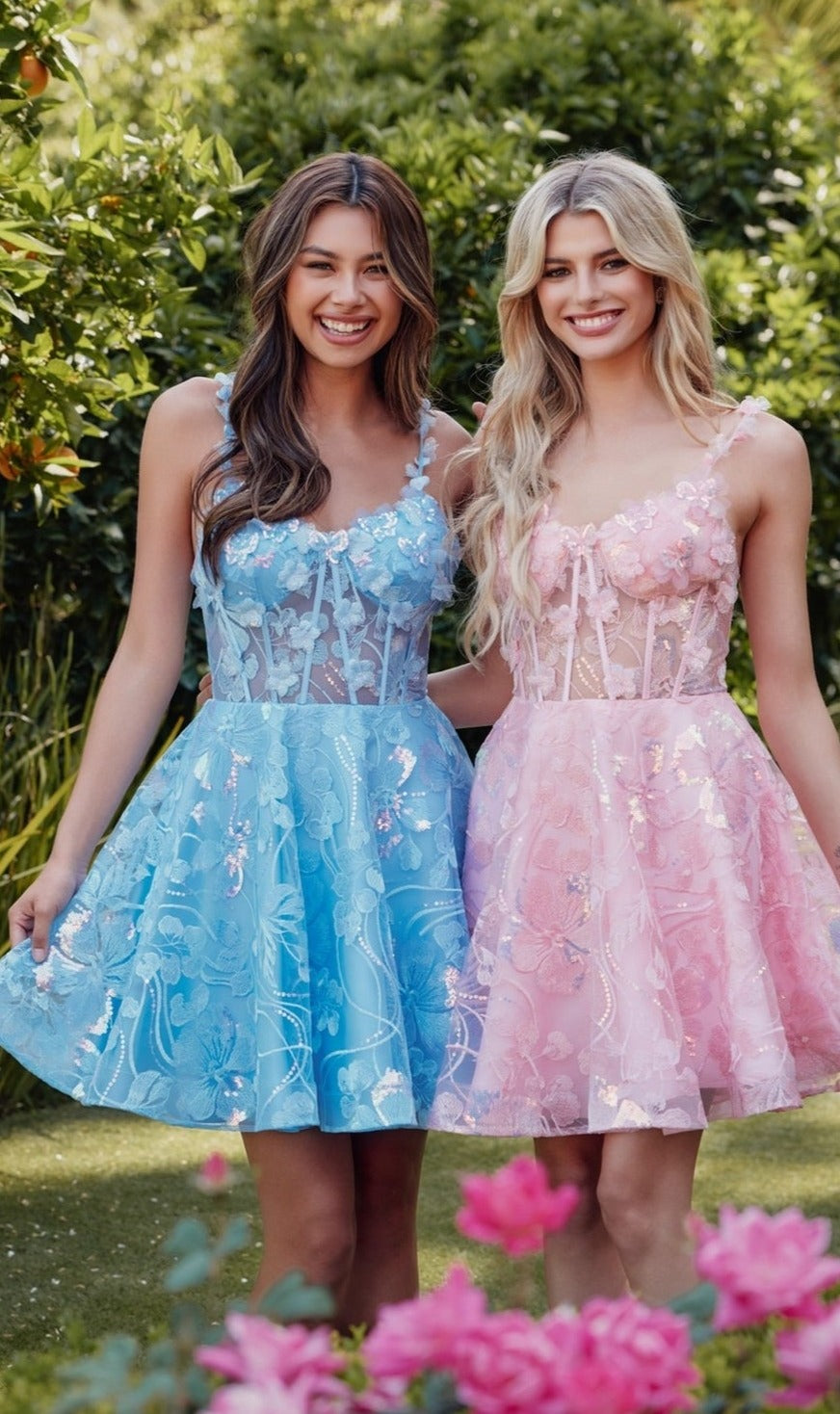 Two young women smile and pose in a garden, one wearing the Sheer-Corset Short Butterfly Hoco Dress JT917A and the other in a pink lace short A-line party dress, surrounded by lush greenery.
