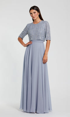A woman wears the Mac Duggal 9180 Short Sleeve Long Formal Dress, featuring a beaded bodice and flowing light blue chiffon skirt, standing against a plain light background.