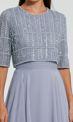 A person wearing the Mac Duggal 9180, a light blue long A-line formal dress with short sleeves and a beaded crop top featuring intricate vertical and horizontal sequin patterns.