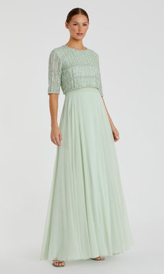 A woman wears the Mac Duggal 9180—a short sleeve, long formal dress with a sequined, beaded crop top and flowing skirt—in light green, standing against a plain white background.