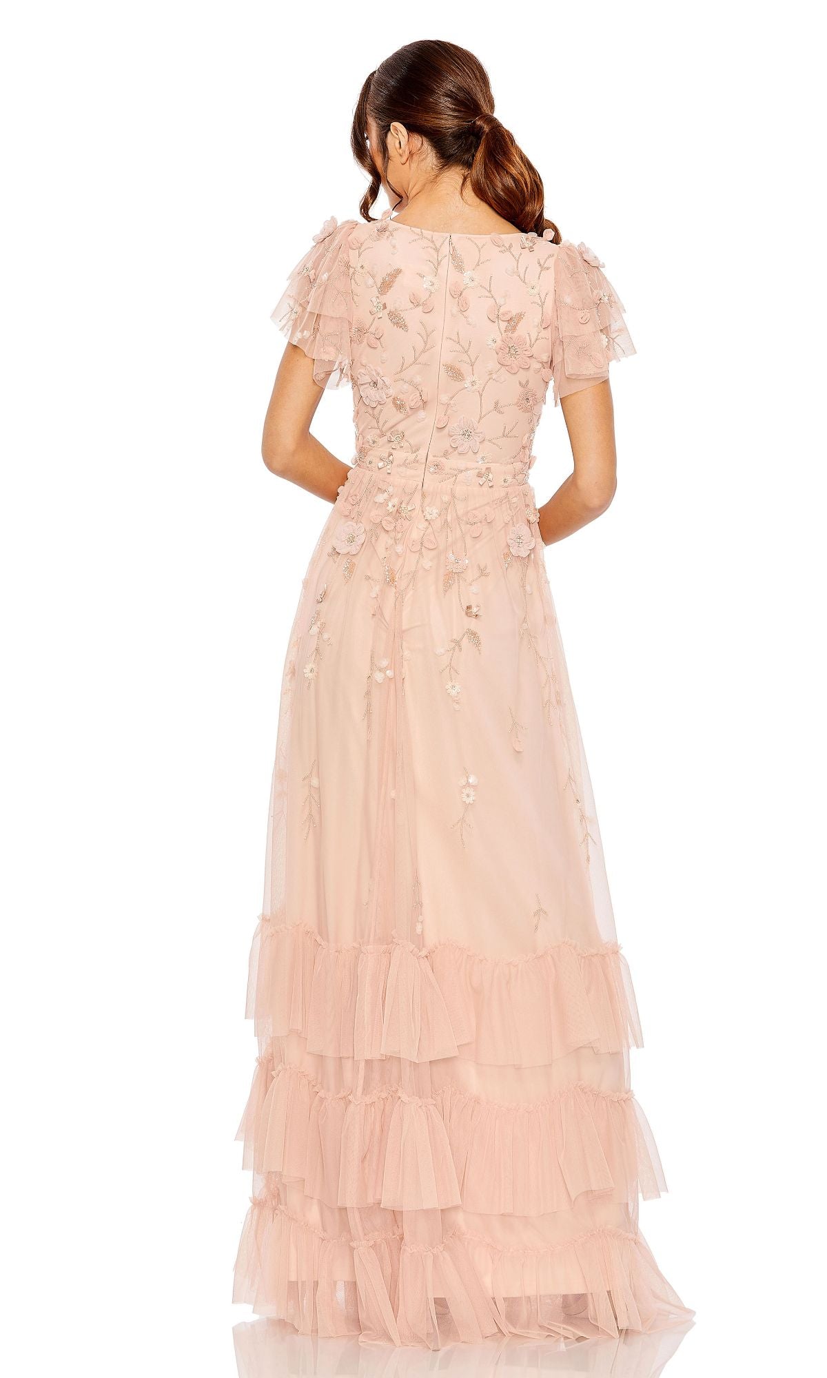 A woman with brown hair in a ponytail is shown from the back wearing the Mac Duggal 9189, a blush pink formal dress with short sleeves, floral embroidery, and layered tulle ruffles.