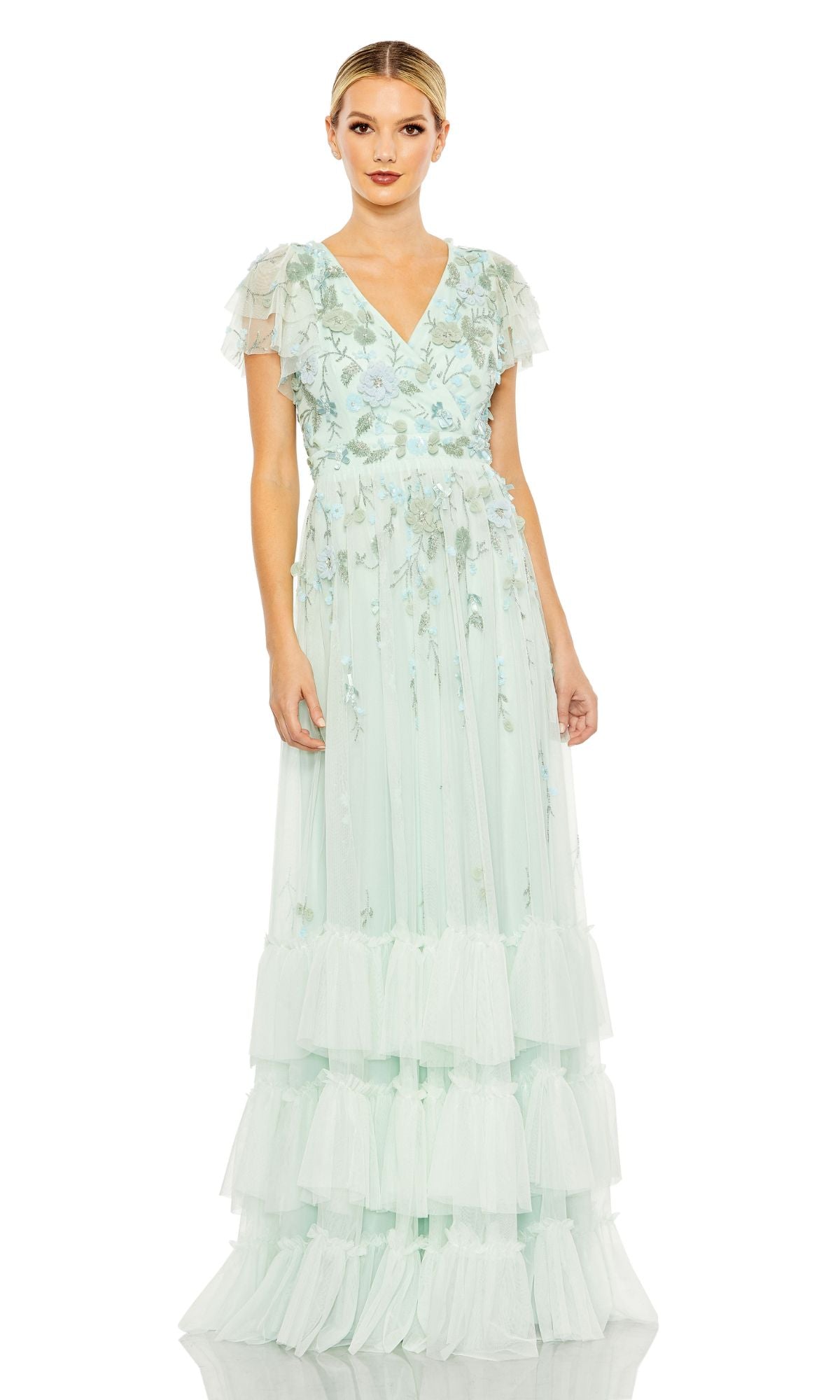 A woman wears the Mac Duggal 9189 Short Sleeve Ruffle Formal Dress, a pastel green floor-length gown with floral embroidery, flutter sleeves, and a layered tulle skirt, standing against a white background.