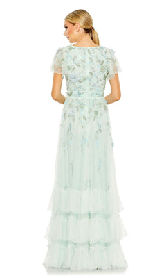A woman faces away, wearing the Mac Duggal 9189 Short Sleeve Ruffle Formal Dress—a floor-length light blue gown with floral print, short sleeves, tiered tulle layers, and delicate embroidery. Her hair is tied back in a low ponytail.