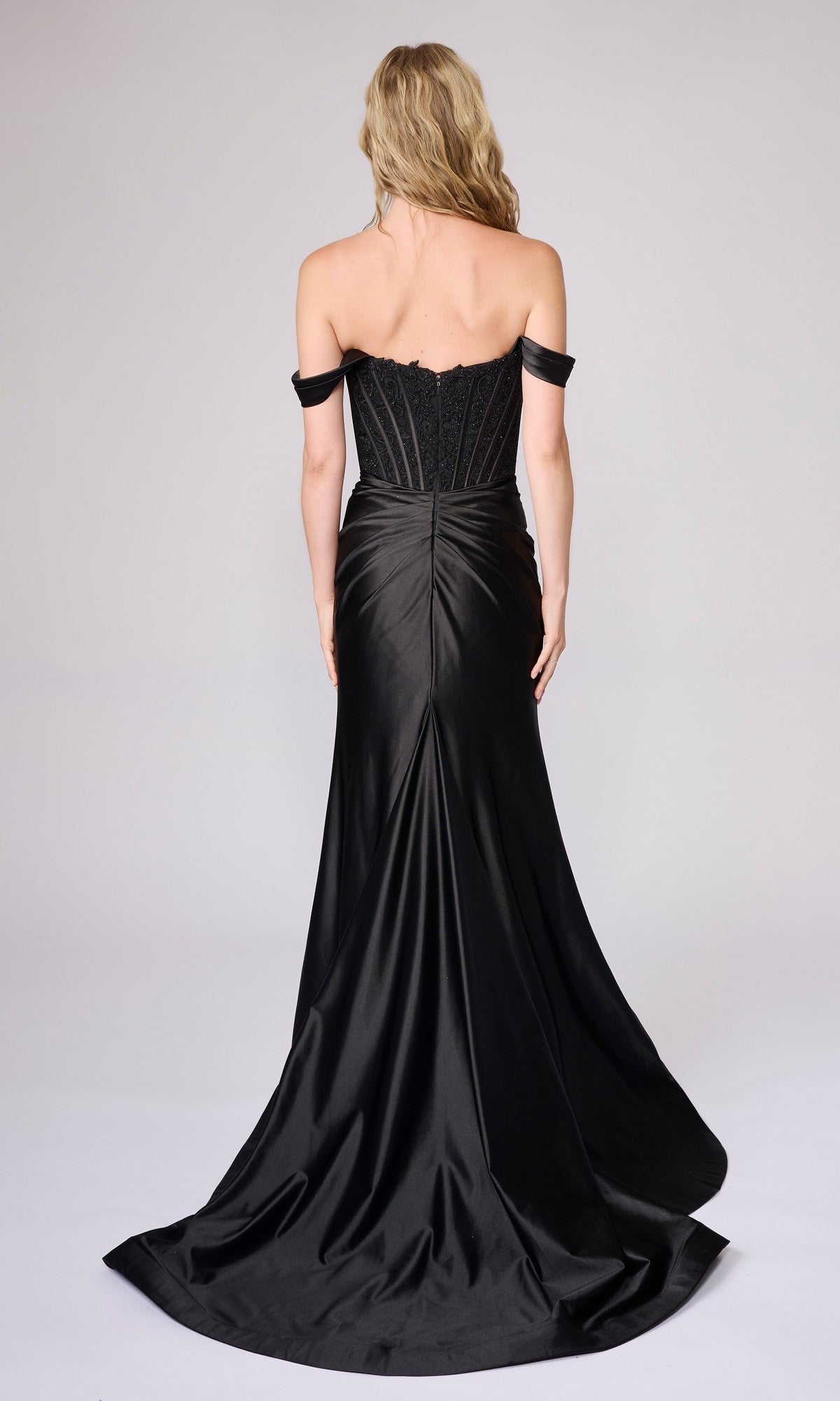 A woman with long blonde hair models the Nina Canacci 9202 long formal dress, featuring an off-the-shoulder embellished corset bodice and flowing floor-length skirt, shown from the back against a plain backdrop.