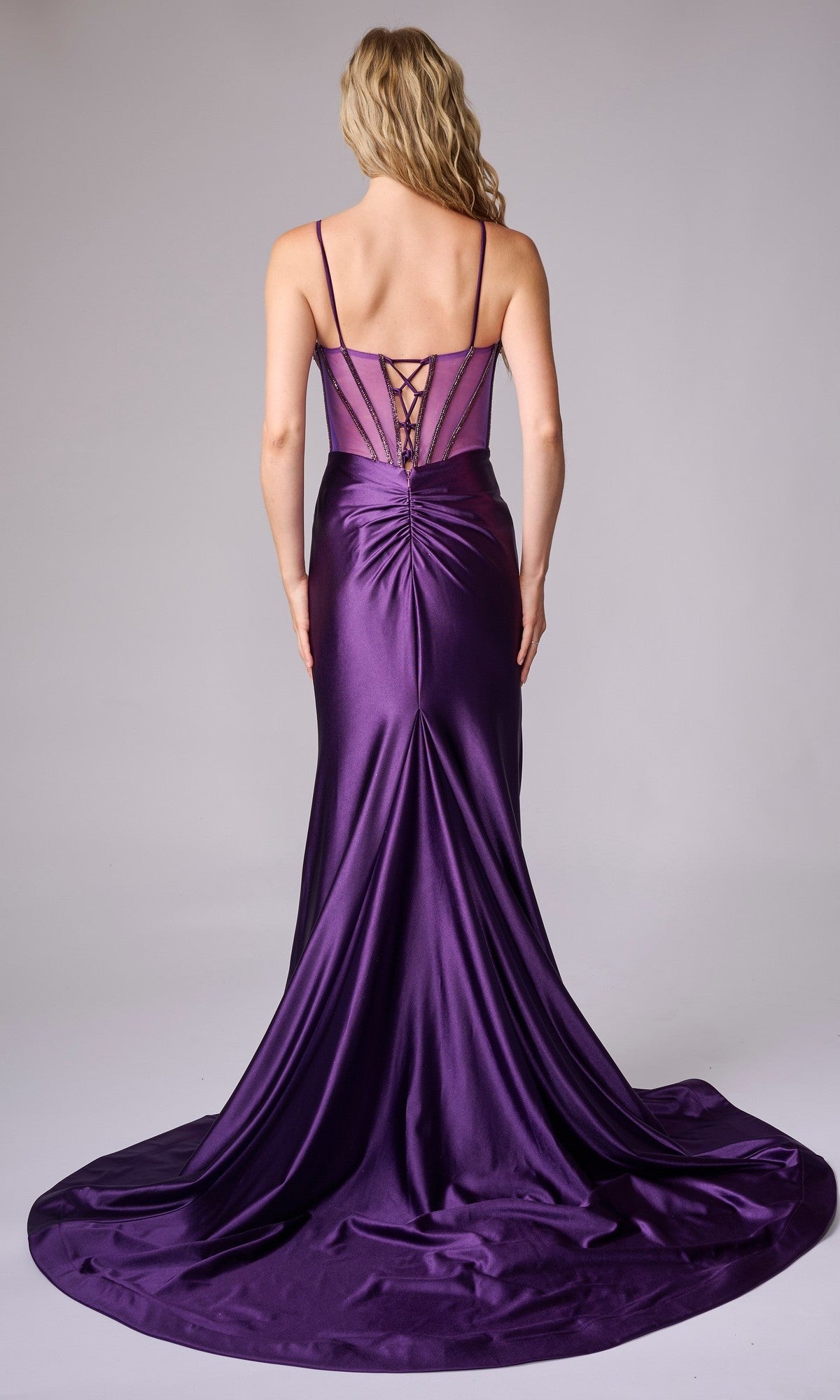 A woman stands against a plain background wearing the Nina Canacci 9206: a floor-length, deep purple formal dress featuring a lace-up back, fitted bodice, and flowing train.