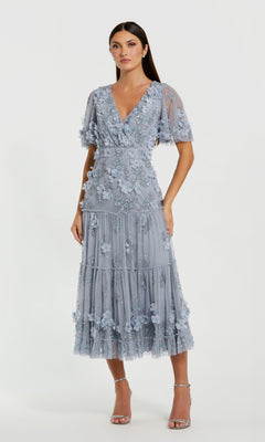 A woman with long dark hair wears the Mac Duggal 9207 A-Line Wedding Guest Dress, featuring a light blue floral design, short sleeves, V-neckline, tiered tea-length skirt, and paired with strappy heels.