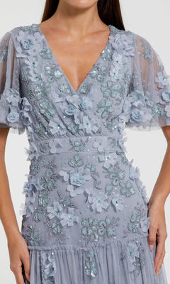 A woman wearing the Mac Duggal 9207 A-Line Wedding Guest Dress, a light blue tea-length gown featuring floral embellishments, embroidered butterfly designs, and short sheer sleeves.