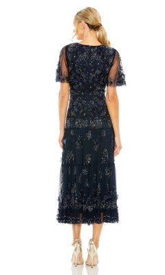A woman stands with her back to the camera, wearing the Mac Duggal 9207 A-Line Wedding Guest Dress in navy blue with floral embellishments, short sheer sleeves, and metallic heels.