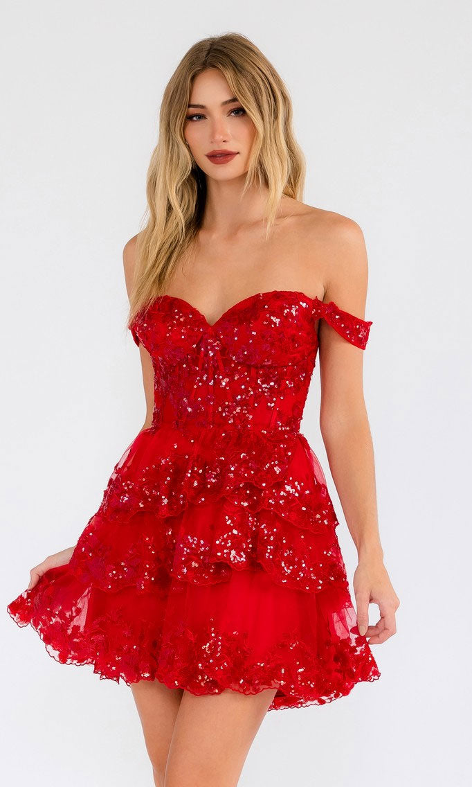 A woman wears the Sequin Sheer Corset Homecoming Dress: Juliet JT920K, a red off-the-shoulder mini dress with sequins and a tiered skirt, standing against a plain light background.