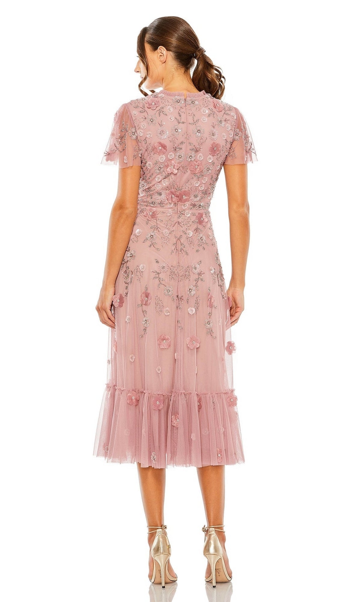 Woman stands with her back to the camera in a pink, floral Mac Duggal 9212 midi wedding-guest dress featuring sheer sleeves and hem, paired with metallic high-heeled sandals.