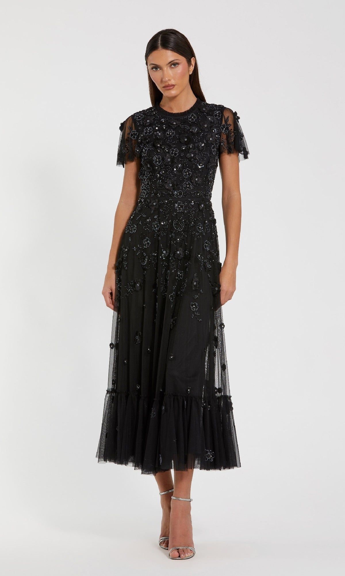 A woman wears the Mac Duggal 9212 Floral Midi Wedding-Guest Dress with short sleeves, floral embellishments, and sheer details, paired with strappy heels, against a plain white background.