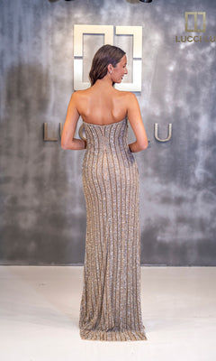 A woman stands facing away in front of a grey Lucci Lu backdrop, highlighting the stunning detail of her floor-length gold and silver Long Prom Dress: Lucci Lu 92145.