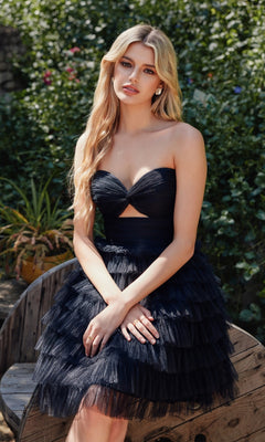 A woman with long blonde hair sits outdoors on a wooden bench, wearing the Ruffled Babydoll Homecoming Dress: Juliet JT921H and earrings, surrounded by greenery.