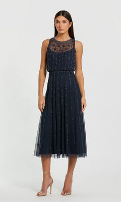 A woman wearing the Mac Duggal 9222 Blue Midi Wedding Guest Dress with sheer embroidered overlay and gold high-heeled sandals stands against a plain white background.