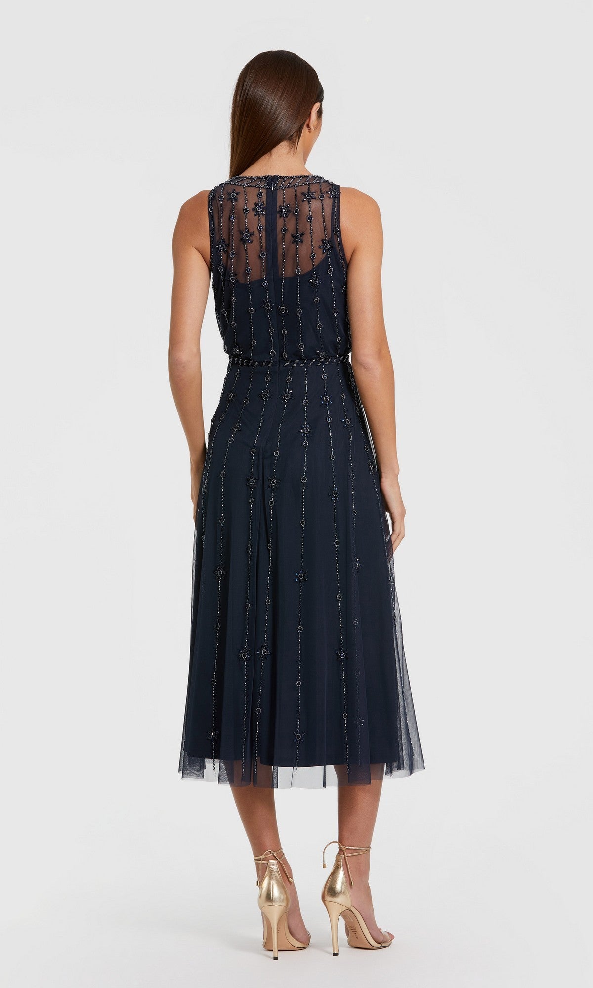 A woman stands facing away in the Mac Duggal 9222 blue midi wedding guest dress, featuring a sleeveless design with a sheer embellished overlay, styled perfectly with gold high-heeled shoes.
