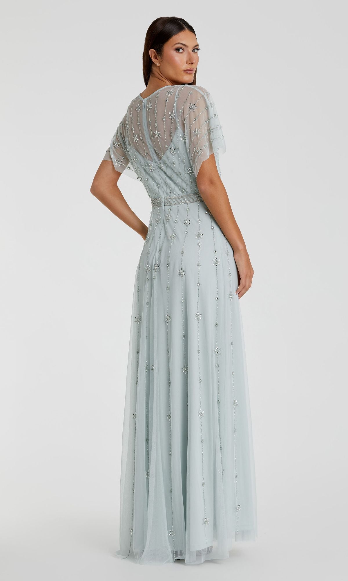 A woman faces backward in a Mac Duggal 9223 light blue short sleeve long formal dress featuring sheer sleeves and delicate bead embellishments, set against a plain white background.