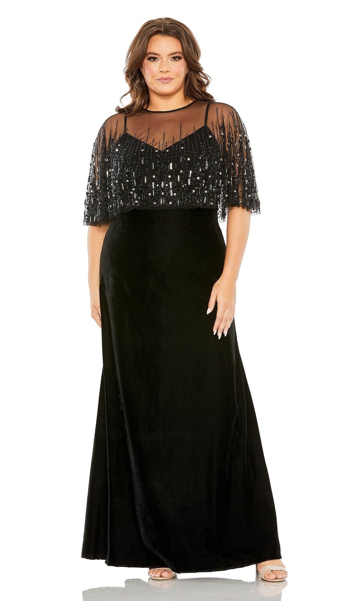 A woman models the Mac Duggal 9228, a long plus-size formal gown in black velvet with a sheer, sequined overlay, standing forward with her hand at her side.