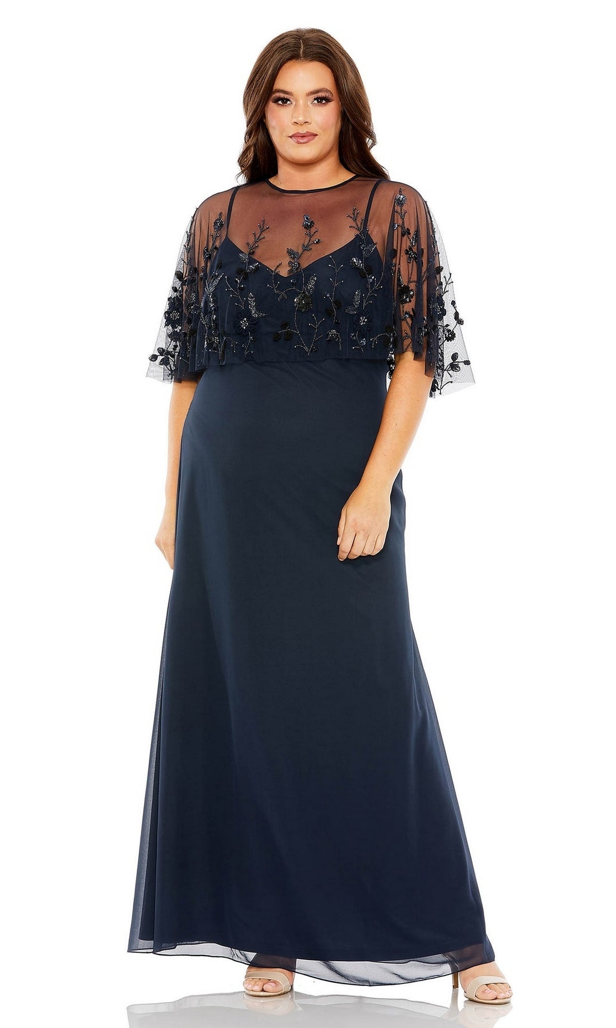 A woman stands against a white background wearing the Long Plus-Size Prom Dress 9230 by Mac Duggal, featuring a floor-length navy blue design with embellishments, sheer embroidered overlay, and short flutter sleeves.