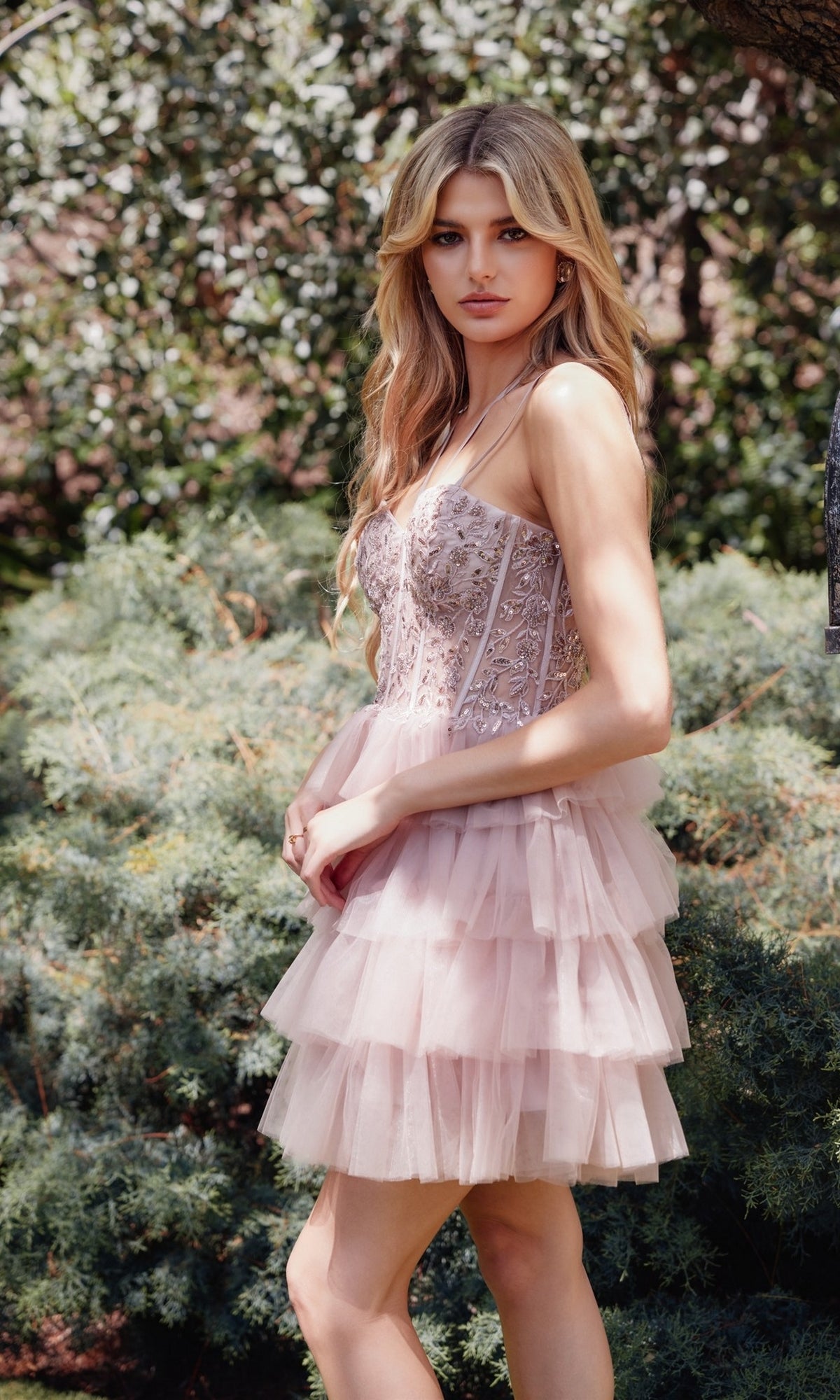 A woman stands outdoors among shrubs and trees, wearing the Sheer-Corset Short Ruffled Hoco Dress JT923S—a pale pink, tiered tulle A-line skirt with a fitted, embellished bodice.