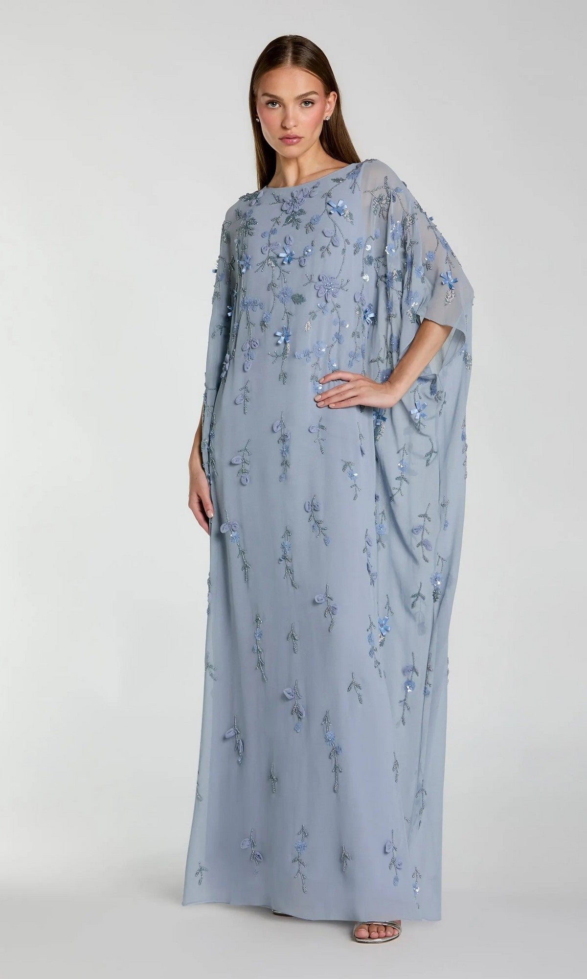 A woman poses against a plain light gray background, wearing the Mac Duggal 9242 Trapeze Embellished Formal Gown—floor-length, loose-fitting, with sheer sleeves and floral embroidery.