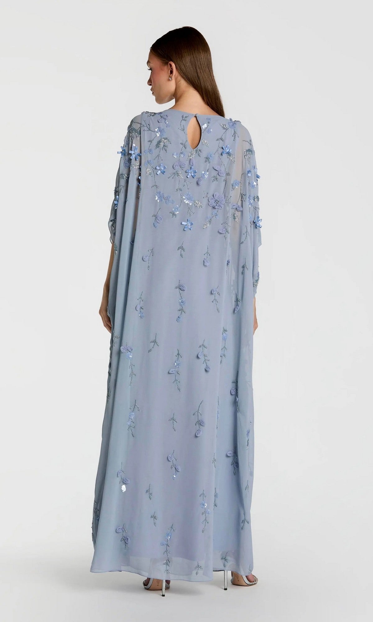 A woman stands with her back to the camera, wearing the Trapeze Embellished Formal Gown: Mac Duggal 9242—a long, light blue dress featuring floral embroidery and sheer sleeves.