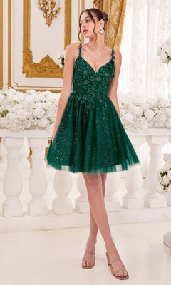 A woman stands indoors wearing the Metallic-Lace Short A-Line Homecoming Dress 9245 in sparkly dark green, paired with nude heels and large earrings. White roses and ornate gold decor provide a glamorous backdrop.