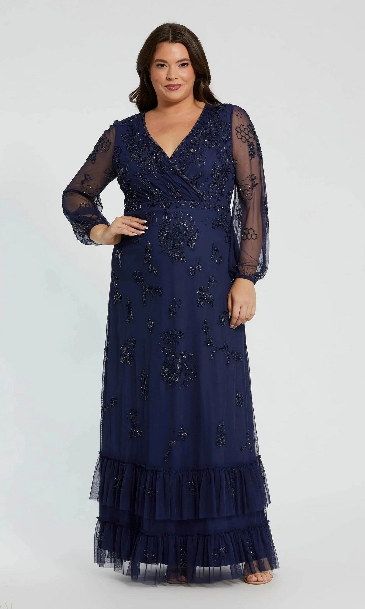 A woman wearing the Mac Duggal 9247, a long plus-size formal dress in midnight, featuring a v-neckline, sheer long sleeves, floral beaded embellishments, and a ruffled hem, stands against a plain light background.