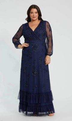 A woman wearing the Mac Duggal 9247, a long plus-size formal dress in midnight, featuring a v-neckline, sheer long sleeves, floral beaded embellishments, and a ruffled hem, stands against a plain light background.