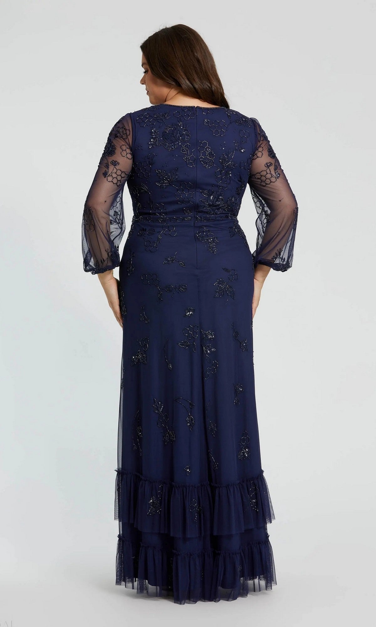 A woman stands with her back to the camera against a plain background, modeling the Long Plus-Size Formal Dress: Mac Duggal 9247—a navy gown featuring sheer embroidered sleeves and a ruffled hem for an elegant formal look.
