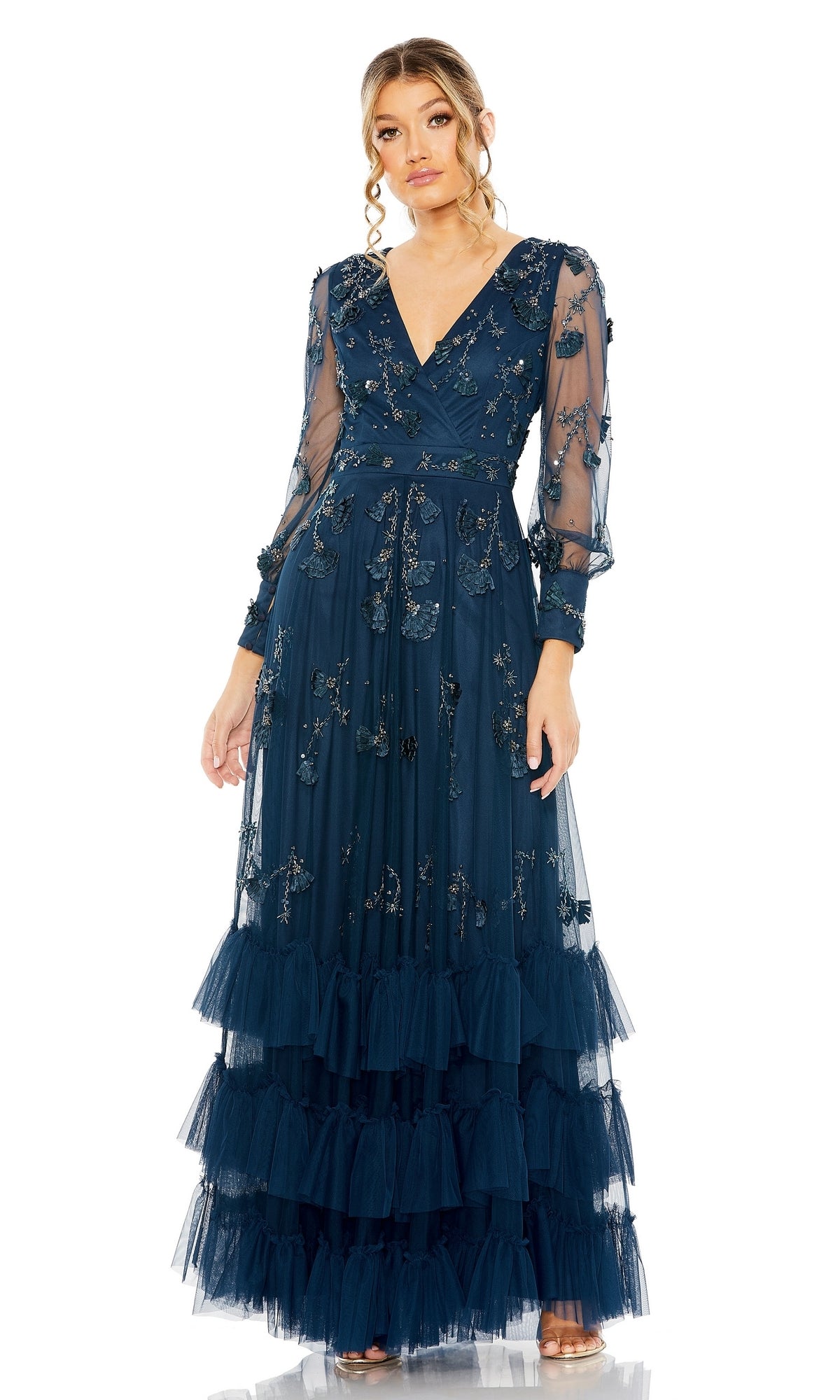 A woman stands against a white background wearing the Mac Duggal 9256 Long Sleeve Blue Formal Dress, featuring sheer embroidered sleeves and a tiered ruffle skirt.