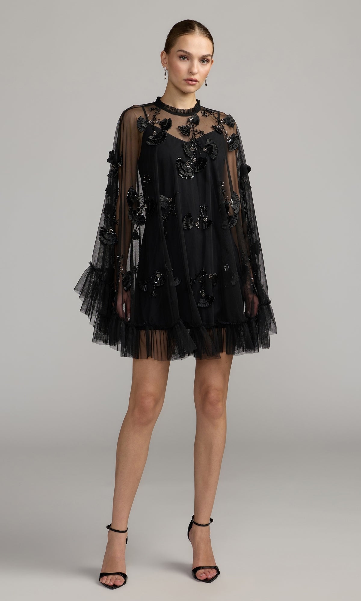 A woman poses in the Mac Duggal 9257 Long Sleeve Short Hoco Dress in black, featuring a sheer embellished overlay and flared sleeves, styled with black ankle-strap heels against a plain background for an effortlessly elegant party look.