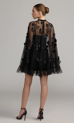 A woman stands facing away against a plain background, wearing the Long Sleeve Short Hoco Dress: Mac Duggal 9257—a short, sheer black ruffled homecoming dress with floral appliques and a ruffled hem—paired with black high heels and earrings.