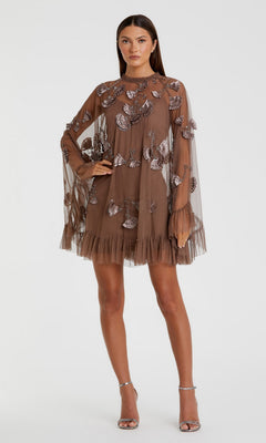 A woman wears the Mac Duggal 9257 Long Sleeve Short Hoco Dress in chocolate, featuring a sheer design with butterfly appliqués and a ruffled hem, paired with silver high-heeled sandals.
