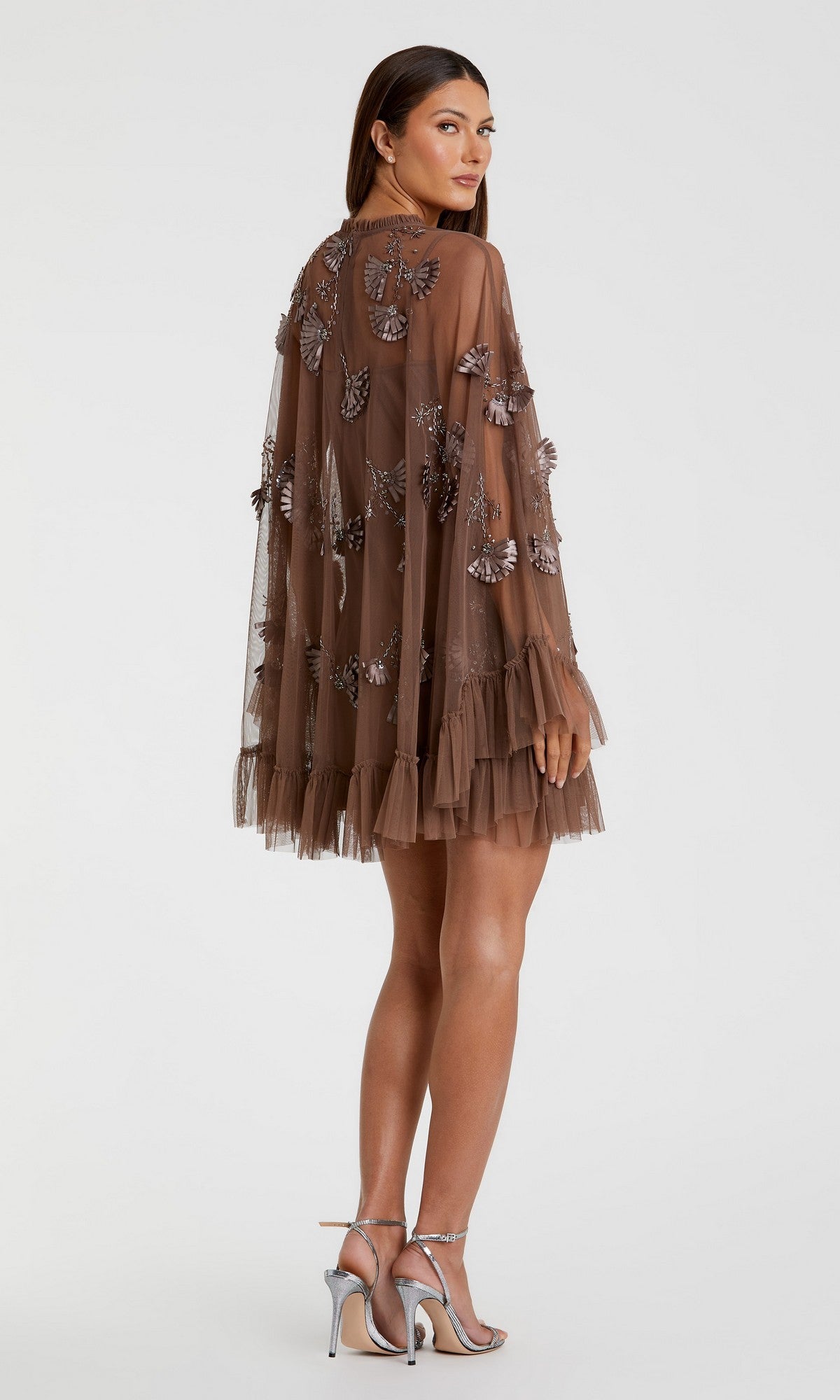 A woman wears the Mac Duggal 9257 Long Sleeve Short Hoco Dress—sheer brown with floral embellishments and a ruffled hem—paired with silver high-heeled sandals, standing against a plain background.