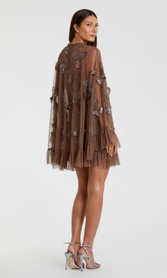 A woman wears the Mac Duggal 9257 Long Sleeve Short Hoco Dress—sheer brown with floral embellishments and a ruffled hem—paired with silver high-heeled sandals, standing against a plain background.