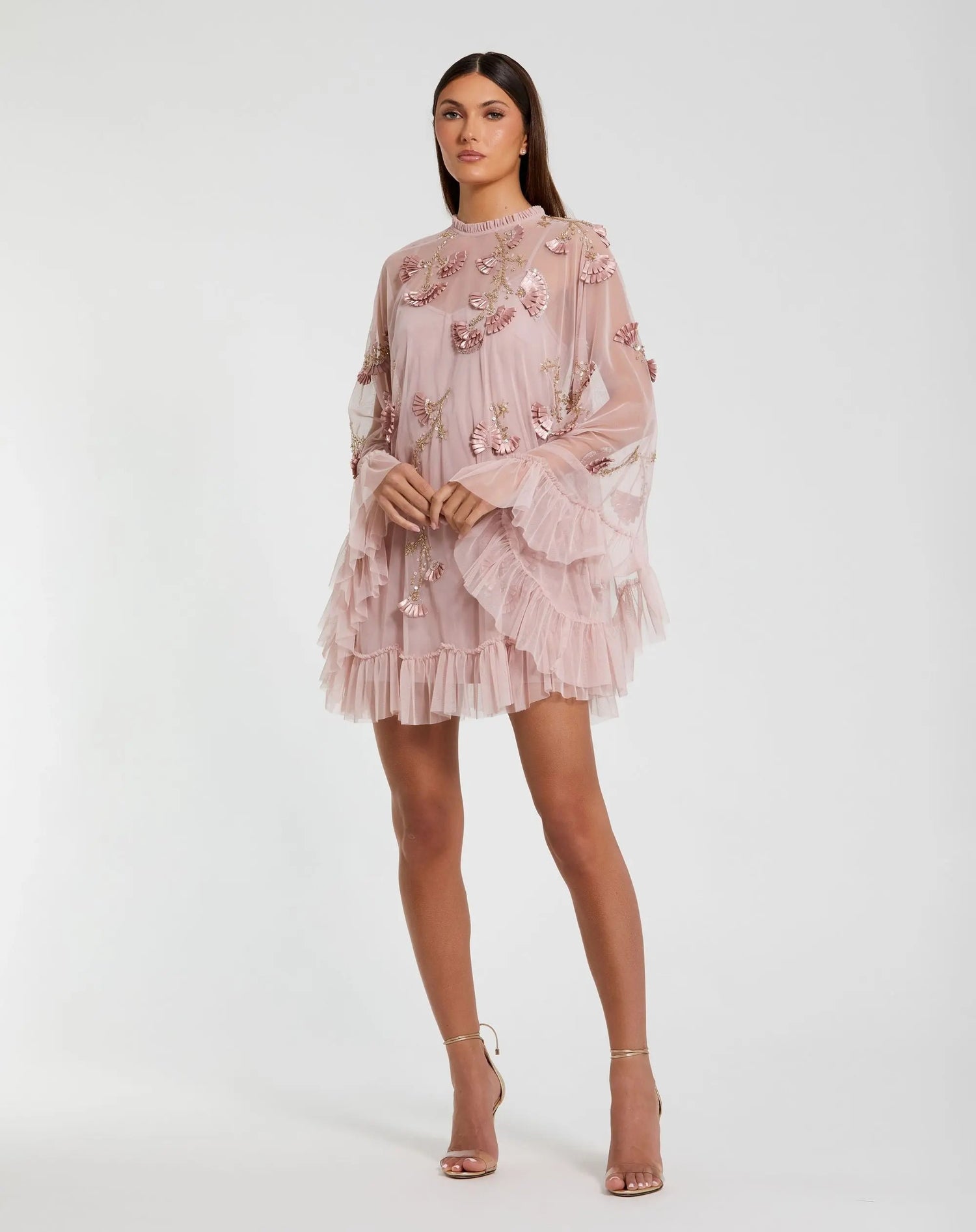 A woman models the Long Sleeve Short Hoco Dress: Mac Duggal 9257, a light pink sheer party dress with floral appliqués, ruffled hem, and high neckline, posing against a plain white background.