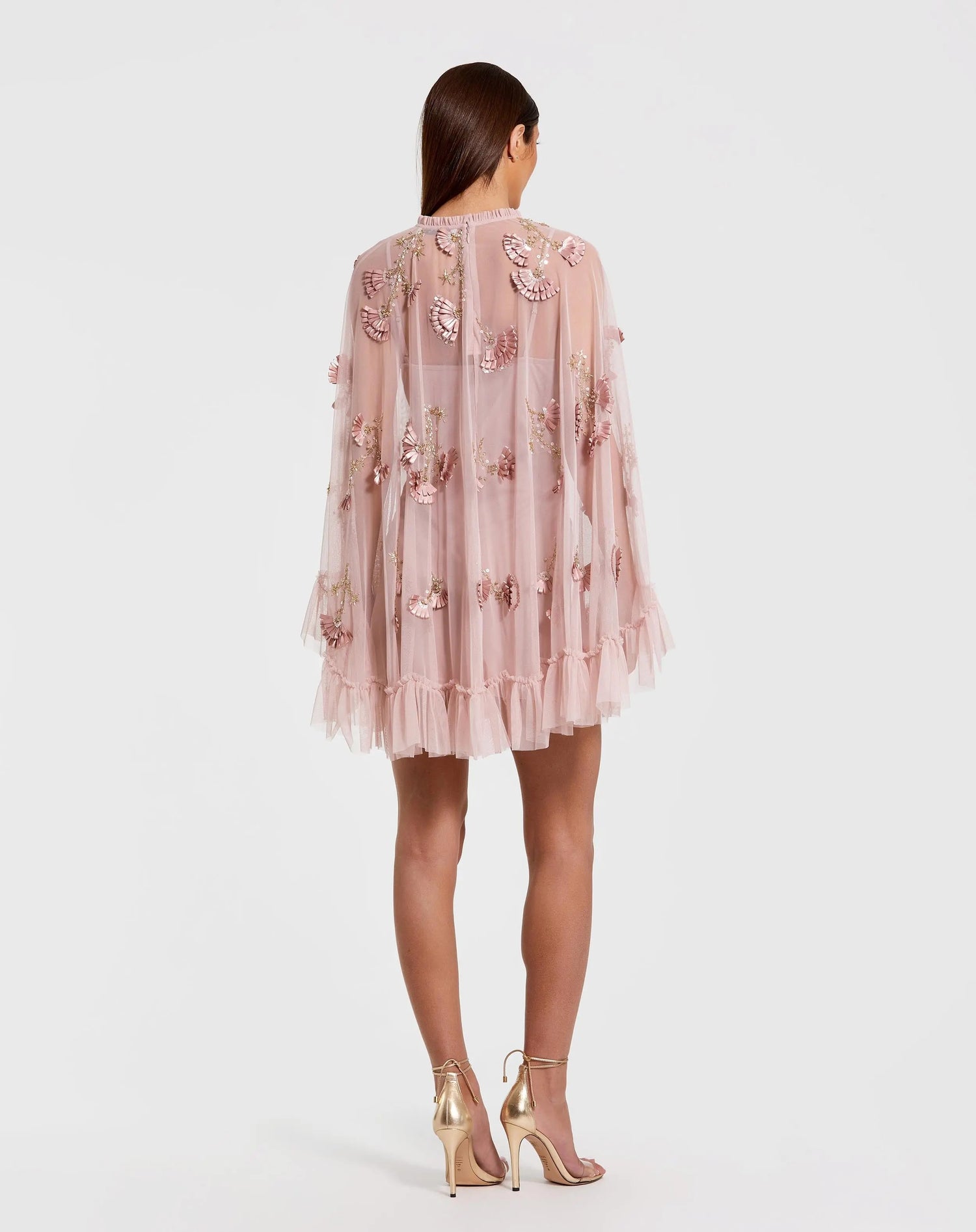 A woman stands facing away, wearing the Mac Duggal 9257 Long Sleeve Short Hoco Dress in light pink with floral appliqué, a sheer cape, and ruffled hem, styled with metallic high-heeled sandals.