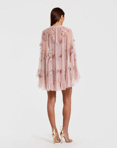 A woman stands facing away, wearing the Mac Duggal 9257 Long Sleeve Short Hoco Dress in light pink with floral appliqué, a sheer cape, and ruffled hem, styled with metallic high-heeled sandals.