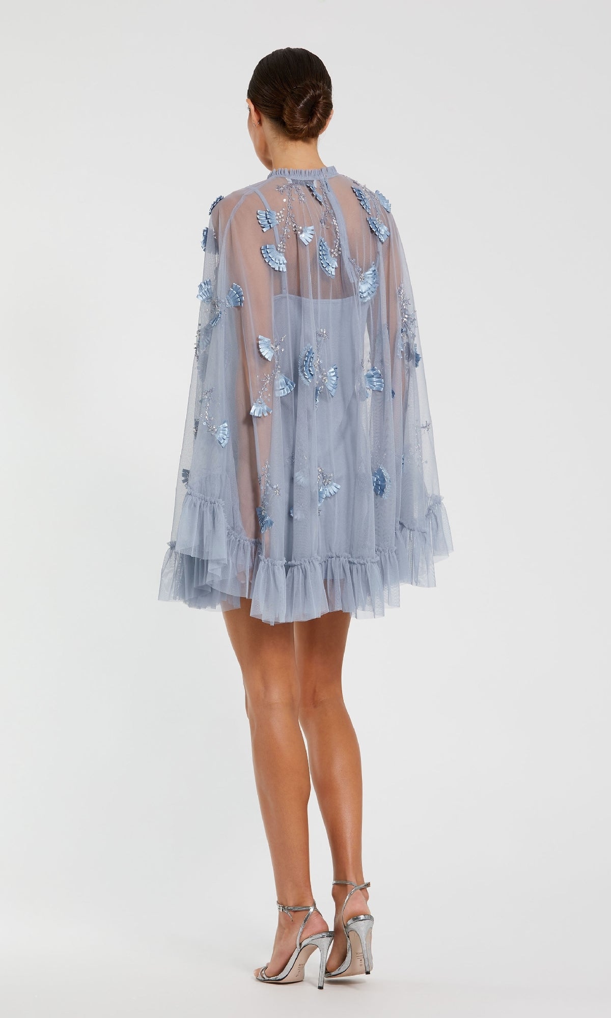 Woman wearing the Mac Duggal 9257 Long Sleeve Short Hoco Dress in sheer light blue with ruffles and butterfly appliqués, standing on a white background and paired with silver high-heeled sandals.